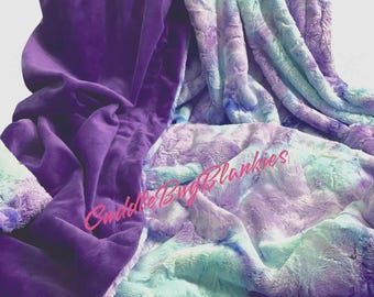 May include: A purple and blue tie-dye plush blanket with a soft, velvety texture on one side. The blanket is folded and draped over a surface. The text "Cuddle Bug Blankies" is visible in the lower left corner of the image.
