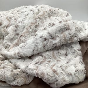 May include: A plush, faux fur blanket in shades of white, cream, and brown. The blanket has a textured, rippled surface, with a solid brown backing. The soft, fluffy material is folded over itself, creating a cozy and inviting appearance.