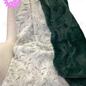 May include: A cozy, plush blanket in shades of white and dark green. The white blanket features a leaf pattern, while the dark green blanket has a solid color. A pink starburst with the word "NEW" is visible.