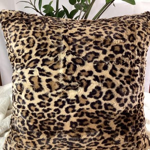 May include: A square decorative pillow with a faux fur leopard print pattern. The pillow features a tan background with dark brown spots. The pillow is placed on a white surface, with green plants in the background.
