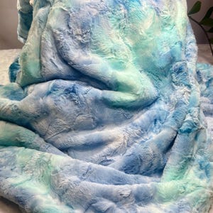 May include: A soft, blue and green tie-dye faux fur throw blanket. The blanket is folded and appears to be made of a plush, fuzzy material.