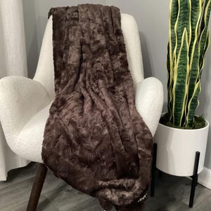 May include: A brown faux fur throw blanket draped over a white armchair. The blanket has a textured appearance and is a rich, dark brown color. A tall snake plant in a white pot is to the right of the chair.
