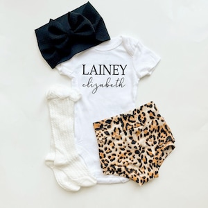 May include: A white baby onesie with the text "LAINEY elizabeth" in black. A black headband with a bow, white socks, and leopard print shorts are also included.