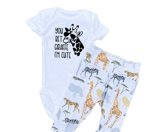 May include: A white baby onesie with the text "You bet giraffe I'm cute" and a black silhouette of a giraffe wearing sunglasses. The onesie is paired with blue pants with a pattern of giraffes, zebras, and other safari animals.