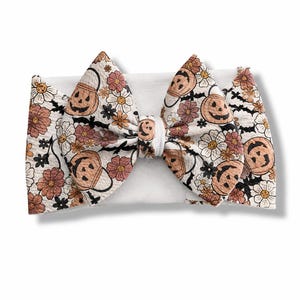 Peut inclure: Un large bandeau blanc avec un grand nœud central. Le tissu présente un motif répétitif de seaux citrouilles orange, de chauves-souris noires et de fleurs variées dans des tons rose, blanc et orange sur un fond blanc.