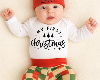 May include: A baby wearing a Christmas outfit, including a white long-sleeved bodysuit with "MY FIRST Christmas" printed on it. The outfit also includes red and green checkered pants and a matching hat with the name "Levi" on it.