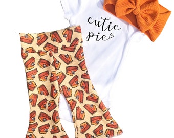Op de afbeelding: Een driedelige baby-outfitset bestaande uit een wit onesie met de tekst "cutie pie" in zwart, een gele broek met wijde pijpen met een pompoentaartprint en een oranje hoofdband.