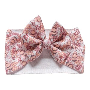 May include: A white headband with a pink and yellow bee print bow. The bow is tied in a knot and the headband is made of a soft, stretchy material.
