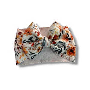 May include: A wide, textured fabric headband featuring a large, knotted bow at the center. The material is off-white with a repeating pattern of orange and yellow flowers, dark green leaves, and small brown sprigs across the entire surface.