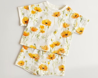 Könnte beinhalten: Ein zweiteiliges Kinder-Outfit, bestehend aus einem kurzärmeligen T-Shirt und passenden Shorts. Das Set ist weiß und mit einem sich wiederholenden Muster aus gelben und orangefarbenen Mohnblumen und grünen Stielen versehen. Der Stoff wirkt weich und leicht.