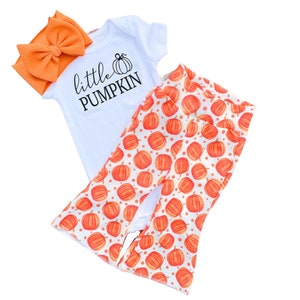 May include: A white onesie with the text "little PUMPKIN" and a small pumpkin graphic. An orange headband with a bow is included. The onesie is paired with white pants with an orange pumpkin print.