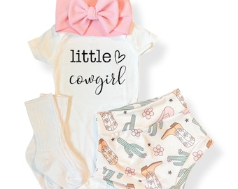 May include: A baby outfit set with a white onesie printed with "little cowgirl", a pink bow headband, white socks, and shorts patterned with cowboy boots, cacti, and flowers. The set is designed for infants.