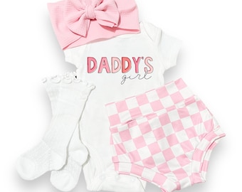 Puede incluir: Un conjunto de ropa para bebé rosa y blanco con un body blanco que dice "Daddy's girl" en letras rosas, bombachos a cuadros rosa y blanco, una diadema con lazo rosa y calcetines blancos.
