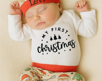 May include: A newborn baby wearing a Christmas-themed outfit. The outfit includes a white onesie with "My First Christmas" printed on it, a matching hat with the name "Levi", and patterned pants with Christmas designs. The outfit has red trim.