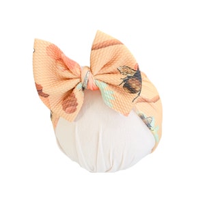 May include: A peach-colored baby headband with a large bow featuring a bee and floral print. The bow is tied in the center and sits atop a soft, cream-colored base. The headband is designed for infants and toddlers.