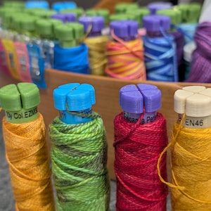 May include: A selection of colourful thread spools, with green, blue, purple, and cream tops. The spools are arranged in a wooden basket. The thread colours include orange, green, red, and yellow. Some spools have text labels.
