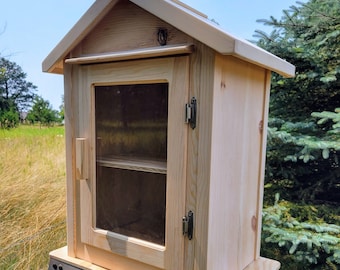 Puede incluir: Un pequeño armario de madera con forma de casa y puerta de cristal transparente. El armario es de madera de color claro y tiene un tejado a dos aguas. Tiene una pequeña balda en el interior y se asegura con pestillos metálicos. El armario está diseñado para uso en exteriores.