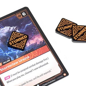 May include: A game card with a wolf illustration and the text "Stormclaw Ursine" is shown with three black and orange "STUNNED" tokens. The card has a "6" in the corner. The tokens are square with the word "STUNNED".