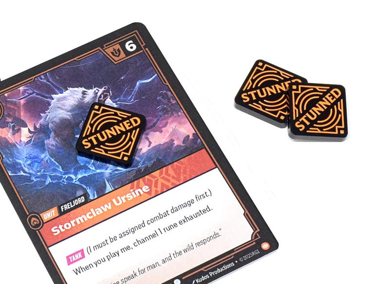 May include: A game card with a wolf illustration and the text "Stormclaw Ursine" is shown with three black and orange "STUNNED" tokens. The card has a "6" in the corner. The tokens are square with the word "STUNNED".