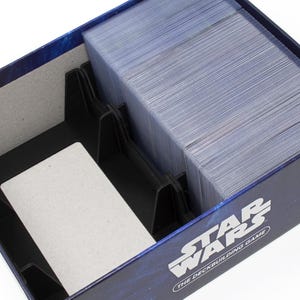 May include: A dark blue Star Wars The Deckbuilding Game box with a black plastic insert featuring dividers. One section contains a large stack of trading cards in clear protective sleeves, while the other section remains empty with a light grey base.