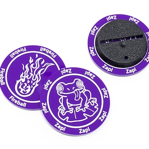 Może przedstawiać: Trzy okrągłe żetony w kolorze fioletowym z białymi wzorami. Jeden żeton przedstawia kulę ognia z płomieniami, drugi żeton przedstawia żabę, a trzeci żeton jest pusty z napisem "Zap!" powtórzonym wokół krawędzi.