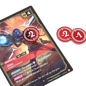 May include: A game card featuring a character illustration with the name "Jinx" and the text "DEMOLITIONIST." Three red game tokens with white numbers 2, 2, and 1 are also present. The card has a black border.