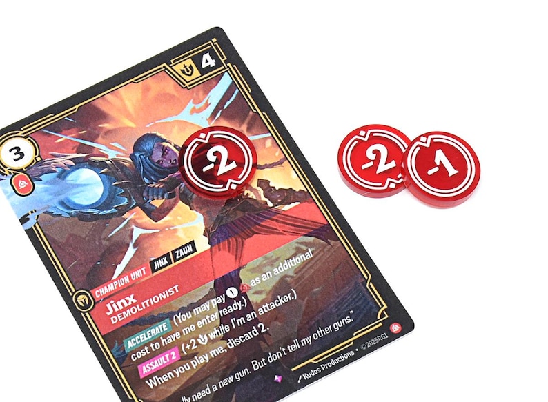 May include: A game card featuring a character illustration with the name "Jinx" and the text "DEMOLITIONIST." Three red game tokens with white numbers 2, 2, and 1 are also present. The card has a black border.