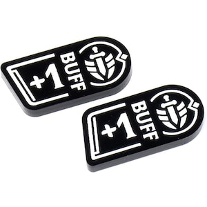 May include: Two black game tokens with white text and symbols. Each token has the words "+1 BUFF" and a shield and sword emblem. The tokens are rectangular with rounded edges.