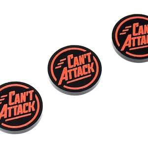 Op de afbeelding: Drie zwarte, ronde tokens met de woorden "Can't Attack" in oranje letters. De tekst staat in een oranje cirkel en de tokens staan op een witte achtergrond. Deze tokens zijn waarschijnlijk voor een bordspel.