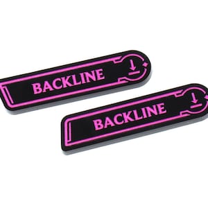Peut inclure: Deux autocollants rectangulaires noirs avec le mot "BACKLINE" en rose vif, souligné de rose. Chaque autocollant a une extrémité arrondie avec un symbole de flèche vers le bas. Les autocollants sont sur fond blanc.