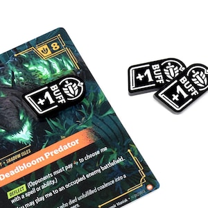 May include: A game card with the text "Deadbloom Predator" and three black and white "+1 BUFF" tokens. The card has a green and orange design with additional text. The tokens are rectangular with rounded corners.