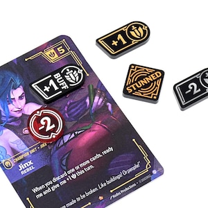 May include: A game card featuring a purple-haired character, Jinx, with various game tokens. The tokens include "+1 BUFF", "-2", "STUNNED", and "+1". The card and tokens are on a white background.