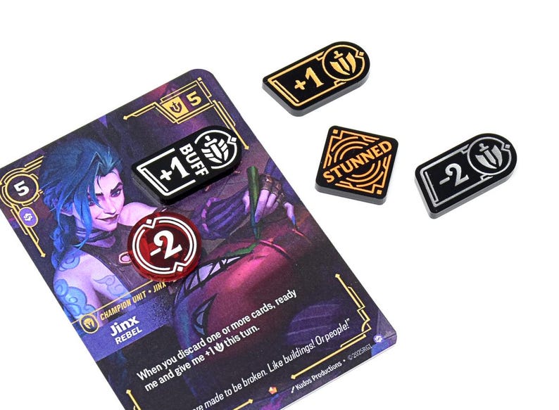 May include: A game card featuring a purple-haired character, Jinx, with various game tokens. The tokens include "+1 BUFF", "-2", "STUNNED", and "+1". The card and tokens are on a white background.