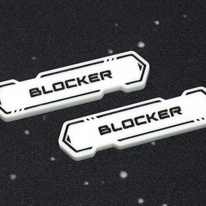 May include: Two white rectangular game pieces with the word "BLOCKER" printed in black. The pieces have a black border and are set against a dark speckled background.