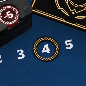 Könnte beinhalten: Nahaufnahme von Spielkomponenten auf einer blauen Oberfläche. Enthält ein schwarzes Tablett mit roten und weißen Spielsteinen und einen goldumrandeten Spielstein mit der Zahl 4. Weiße Zahlen 2, 3, 5 und 6 sind ebenfalls sichtbar.