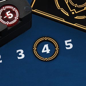 May include: Close-up of game components on a blue surface. Includes a black tray with red and white tokens, and a gold-rimmed token with the number 4. White numbers 2, 3, 5, and 6 are also visible.