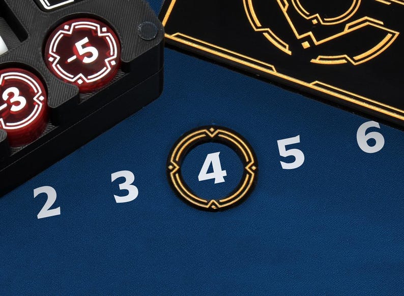 May include: Close-up of game components on a blue surface. Includes a black tray with red and white tokens, and a gold-rimmed token with the number 4. White numbers 2, 3, 5, and 6 are also visible.