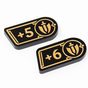 Op de afbeelding: Twee zwarte spel fiches met gouden details. Eén fiche toont "+5" en de andere "+6". Elk fiche heeft een gouden schild- en zwaardontwerp. De fiches zijn rechthoekig met afgeronde randen.
