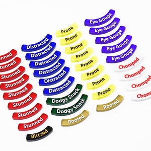 May include: A selection of curved game tokens in a variety of colours, including red, blue, yellow, green, white, black, and gold. Each token has a different word printed on it, such as "Stunned", "Distracted", "Prone", "Eye Gouge", "Dodgy Snack", "Rooted", "Blitzed", and "Chomped".