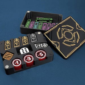 May include: Black game organiser with compartments for tokens, dice, and cards. Includes tokens with +1, +3, -1, and -5 markings, a white die, and a black card with a gold design. The organiser has the text "STUNNED" and "DEATHKNELL".