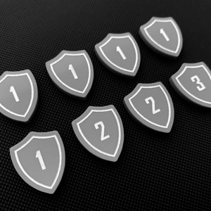 May include: Eight gray shield-shaped tokens with white numbers on a black background. The numbers are 1, 1, 1, 1, 2, 2, and 3.