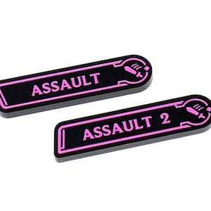 Könnte beinhalten: Zwei schwarze, rechteckige Spielmarken mit abgerundeten Enden. Das Wort "ASSAULT" ist in Pink auf dem oberen Token und "ASSAULT 2" auf dem unteren Token aufgedruckt. Jedes Token hat einen pinkfarbenen Rand und eine kleine Grafik.