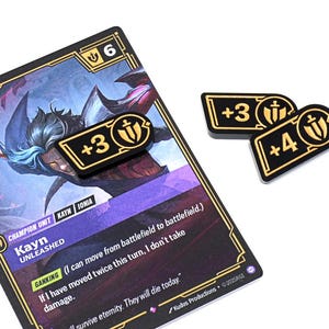 May include: A trading card featuring a character illustration with the text "Kayn Unleashed." Three black and gold game tokens with "+3" and "+4" are placed near the card. The card has a purple and black design.