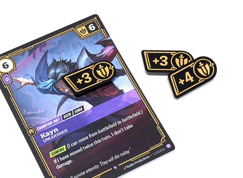 May include: A trading card featuring a character illustration with the text "Kayn Unleashed." Three black and gold game tokens with "+3" and "+4" are placed near the card. The card has a purple and black design.