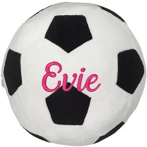 May include: A plush soccer ball in white and black, featuring the name "Evie" embroidered in pink. The ball has a classic pentagon design and is likely a decorative or children's item.