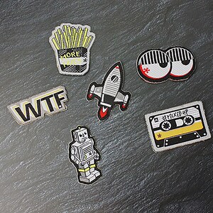 Può includere: Un set di sei toppe da stirare con una varietà di disegni, tra cui un razzo, un robot, una cassetta, un paio di occhi, patatine fritte e le lettere "WTF".
