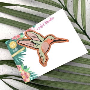Könnte beinhalten: Ein glitzernder Kolibri-Aufnäher in Rosa und Grün mit dem Text "* perfect Paradise" auf weißem Hintergrund.