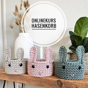 May include: Three crocheted bunny-shaped baskets in gray, pink, and green. The baskets have black eyes and a pink nose. The baskets are sitting on a wooden shelf with the text "ONLINEKURS HASENKORB" in a white circle above them.