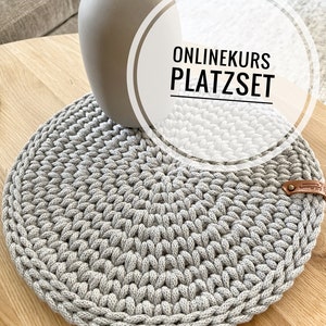 Pode incluir: Um tapete de crochê redondo cinza com uma etiqueta de couro. O tapete está sobre uma mesa de madeira com um vaso e flores secas.