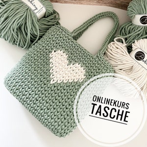 Pode incluir: Uma bolsa de mão de crochê verde com um design de coração branco. A bolsa é feita com corda de algodão grossa e tem um estilo simples e minimalista. O texto "ONLINEKURS TASCHE" é visível em um círculo branco.